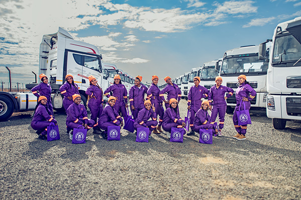 16 women truck-drivers posing in purple outfits in front of a fleet of trucks.