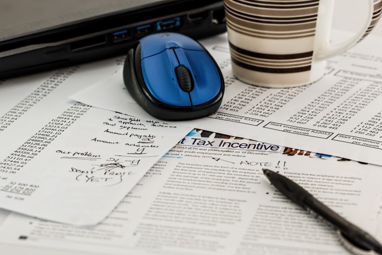 A coffee cup, pen and wireless mouse resting on a bunch of papers on a desk.