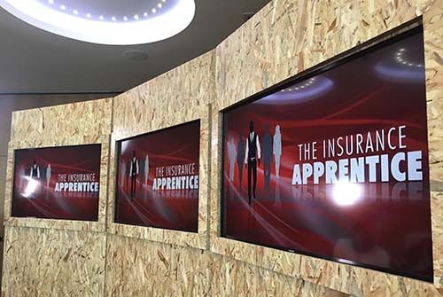 A wall with 3 large screen displaying "The Insurance Apprentice" title.